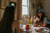 Two women sit at a dining table with lit candles, drinks, and plates of food, smiling and conversing in a cozy, wood-paneled room.