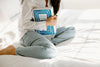 Person sitting cross-legged on a bed, holding a large cookbook titled Greece: The Cookbook, dressed in light jeans and a white shirt.