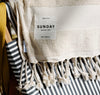 A beige towel with fringe and a Sunday Supply Co. label is draped over a black and white striped bag.