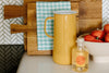 A yellow pitcher, a bottle of Brightland vinegar, a wooden cutting board with a checkered cloth, and a bowl of pomegranates on a kitchen counter.