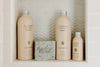Four Flamingo Estate Euphoria bath products—body wash, body lotion, a bar soap, and body oil—are arranged on a tiled shower shelf with a herringbone pattern.