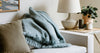 A light blue fuzzy blanket is draped over a white sofa next to a beige pillow, with a lamp, plant, and candles on a wooden side table.