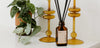 A brown glass diffuser bottle with black reeds sits on a white surface beside two amber glass candleholders and a potted green plant.