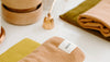 Folded peach and green towels on a white surface with a small gold bell and beige containers in the background. A visible label on one towel reads seadate.