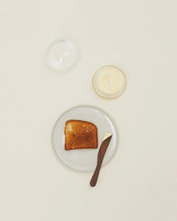 A slice of toasted bread with butter sits on a plate beside a Hawkins New York Simple Glass Butter Keeper and an empty glass bowl, all arranged on a light-colored surface.