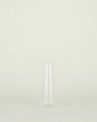The Aurora Slim Drop Vase in Clear by Hawkins New York, featuring handblown glass with a narrow neck and wide base, is centered against a plain, light beige background.