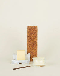 A tall crispbread stands upright behind an unwrapped butter stick, the Simple Glass Butter Keeper by Hawkins New York, and a brown butter knife, all arranged on a plain light background.