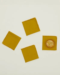 A set of four Essential Cocktail Napkins in Mustard from Hawkins New York, made of 100% cotton with frayed edges, are displayed on a light surface, one napkin elegantly holding a glass of iced beverage.