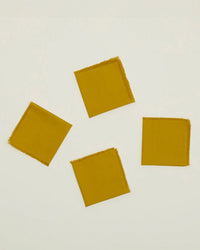The Essential Cocktail Napkins in Mustard by Hawkins New York, a set of 4 made from 100% cotton with frayed edges, are shown loosely scattered on a light background.
