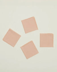 A set of four Essential Cocktail Napkins in Blush by Hawkins New York, made from 100% cotton with frayed edges, are arranged neatly on a plain, light background.