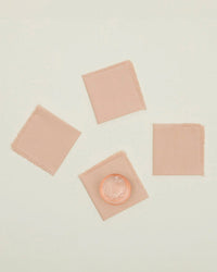 A set of four blush Essential Cocktail Napkins by Hawkins New York, made from 100% cotton, is arranged on a pale surface with a glass of water and ice on the bottom left napkin.