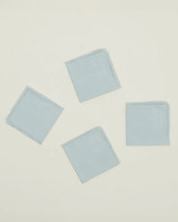 A set of four Essential Cocktail Napkins in Sky by Hawkins New York, made from 100% cotton with frayed edges, are arranged loosely on a plain off-white background.