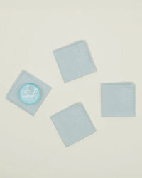 The Hawkins New York Essential Cocktail Napkins in Sky (set of 4) are arranged on a light surface, with one napkin holding a glass of sky-blue drink while the other three remain empty and evenly spaced.