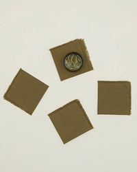 The Essential Cocktail Napkins in Olive by Hawkins New York—a set of four square cotton napkins with frayed edges—are shown, one holding a dark saucer of green olives. Ideal for gatherings or cocktails.