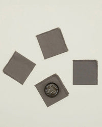 A set of four dark grey Essential Cocktail Napkins by Hawkins New York is displayed on a light surface, with one napkin holding a round object with a face design—ideal for adding style to your cocktail hour.