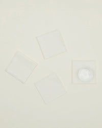 A set of four Essential Cocktail Napkins in Ivory by Hawkins New York is arranged on a light beige background, and one napkin is topped with a small round clear plastic container.