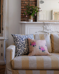 A beige striped sofa with two decorative pillows, including the Soil to Studio Babita Linen Pillow in Old Rose (18x18), sits before a white mantle and exposed brick wall.