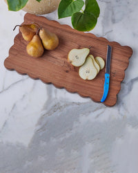 The etúHOME New Scalloped Cutting Board, Medium Rectangle Plank, displays fresh pears and a blue-handled serrated knife on marble with green leaves—an ideal choice for stylish modern kitchen decor.