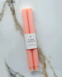 Two Coral Pink Taper Candles | Fluted by BOTANICA, bundled with a white label reading "Taper Candles," rest on veined white marble—elegant hand-dyed decor for any occasion.