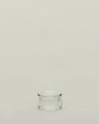 A small, empty Simple Glass Butter Keeper by Hawkins New York with a lid sits centered on a plain, light beige background, evoking minimalist style and leaving ample empty space above.