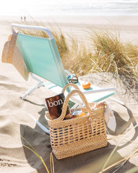 A light blue beach chair with a sun hat sits on sandy dunes near tall grass, facing the ocean. In the foreground, the caminito Canasta Mini Bag in Natural holds a book, sunglasses, a drink, and a napkin.
