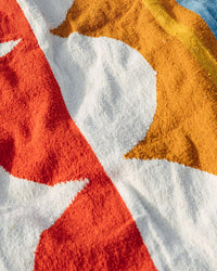 A close-up of the Luna Paseo - Blanket Roll by caminito, a colorful, lightweight outdoor blanket featuring a bold abstract pattern in white, red, orange, and blue. The textured fabric stands out clearly in the sunlight.