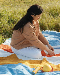 A woman with long dark hair sits on the colorful, lightweight Luna Paseo - Blanket Roll by caminito in a grassy field, wearing a tan jacket and white pants. In front of her are a can, a yellow round object, and an orange-yellow toy.