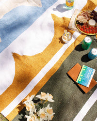 A vibrant picnic unfolds on the Luna Lago - Blanket Roll by caminito, featuring drinks, snacks, a book, notebook, fresh flowers in a jar, and soda cans—sunlit and secured with a handmade leather strap around the lightweight blanket.