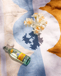A glass bottle, empty plastic cup, and a white flower vase rest on the colorful Luna Lago - Blanket Roll by caminito, featuring blue, white, and orange patterns that cast soft sunlight shadows.
