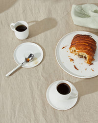 A partially eaten chocolate croissant sits on a plate next to two Simple Porcelain Espresso Cups & Saucers by Hawkins New York, with a spoon on a saucer and a folded napkin atop a beige tablecloth.