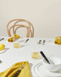 A minimalist dining table set with Hawkins New York Simple Porcelain Salad Plates (set of 4), white plates, yellow napkins, silverware, glasses with light drinks, a wooden chair in the background, and a lemon on a small plate to the left.