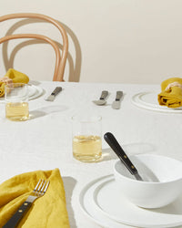 A minimal table setting features Hawkins New York Simple Porcelain Large Bowls, white plates, black-handled cutlery, yellow cloth napkins, and light drinks on a white tablecloth, with wooden chairs in the background.