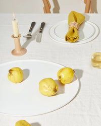 A minimal table setting with a white tablecloth, Hawkins New York’s Simple Porcelain Serving Platter holding three yellow quinces, a drink, candle, cutlery, and a plate with a mustard yellow napkin—all in bright lighting.