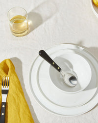 A table setting with a Hawkins New York Simple Porcelain Dinner Plate, matching bowl, a spoon in the bowl, a fork on a yellow napkin, and a glass of light-colored drink atop a crisp white tablecloth.