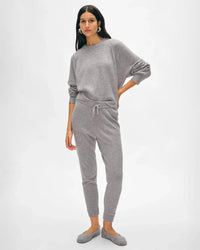 A woman with long dark hair poses calmly in the White + Warren Cashmere Jogger in Grey Heather—a cozy loungewear set with a relaxed fit, long sleeves, drawstring waist pants, and gray slippers, all against a plain background.
