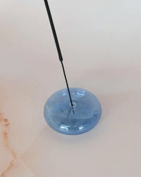 A thin black incense stick stands in the center of BOTANICA’s Bubble Glass Incense Holder - Blue Moon Glass, which rests on a light-colored surface.
