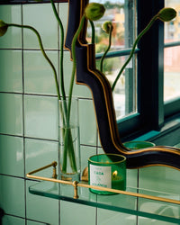 A ROEN Casa Polanco Candle and a tall glass vase with long-stemmed flowers rest on a glass shelf before a mirror, set against glossy green-tiled walls and a window, filling the space with premium fragrance oils.
