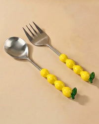 The Casa Amarosa Salad Servers with Lemon Handles S/2 include a metal spoon and fork, each with a lemon-shaped yellow handle and green leaf at the end, displayed on a light beige surface.