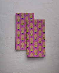 Two folded Kesya Table Napkins S/4 in Fuchsia from Soil to Studio, featuring a yellow and peach pattern with green accents, rest on a textured white surface—ideal for complementing Soil to Studio’s handcrafted decor.