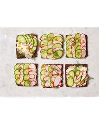 Six pieces of rectangular toast with cream cheese, cucumbers, radishes, carrots, and fresh herbs are arranged on marble—perfect for self-care meals or snacking dinners inspired by Snacking Dinners from Chronicle Books.