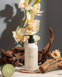 A white pump bottle labeled Brooklyn Candle Studio Maui Liquid Hand + Body Wash is shown with driftwood, orchids, halved lime, and a small flower on a neutral background with soft lighting—highlighting its organic ingredients.