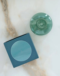 A round teal Bubble Glass Incense Holder by BOTANICA and a matching blue box with a white circle on the lid rest on marble with beige and tan veining.
