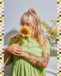 A woman with long blonde hair and glasses, wearing a light green dress, holds a sunflower at Baske's Thanksgiving - Floral Workshop, standing before a white wall and flowers and showcasing the elegance of floral design.