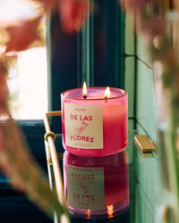 A pink ROEN De Las Flores Candle with two lit wicks sits on a reflective surface near a window, surrounded by blurred floral elements inspired by the Mexico City flower market.