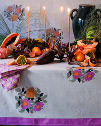 A table draped with a floral cloth displays assorted fruits, foliage, five candles, and a dark ceramic jug. Soil to Studio Kesya Table Napkins in Fuchsia and their linen cushion covers add handcrafted charm amid grasses and floral decor.