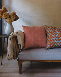 A cozy sofa with wooden legs holds two patterned pillows—one featuring Soil to Studio's Rekha Linen Pillow (18 x 18 in, Rojo), the other blue with orange designs. A beige throw is draped nearby, next to a white pitcher of dried flowers on a small table.