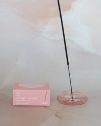 A BOTANICA Bubble Glass Incense Holder in Coral Pink holds a lit stick beside its branded box, set against a soft, neutral background.