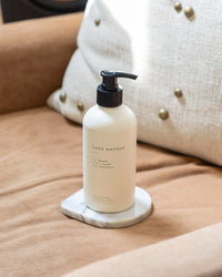 A white pump bottle of Brooklyn Candle Studio Love Potion Liquid Hand + Body Wash rests on a small marble tray atop a tan upholstered chair with a cream, brass-studded cushion in the background.