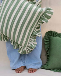 A barefoot person in loose light blue jeans holds the Casa Amarosa Striped Green Ruffle 16x16in Pillow; another green-ruffled, 100% cotton pillow from the same brand is nearby.