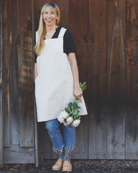 A smiling woman stands before a wooden wall, wearing The Floral Society's Canvas Workshop Apron in Natural over a black shirt and blue jeans. She holds pale pink peonies and finishes the look with clogs and patterned socks.